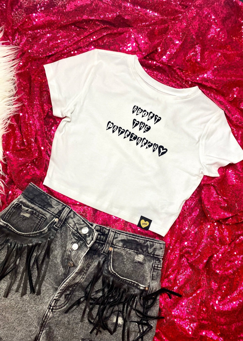 abort the patriarchy crop top on a hot pink sequin background with black denim shorts with leather fringe