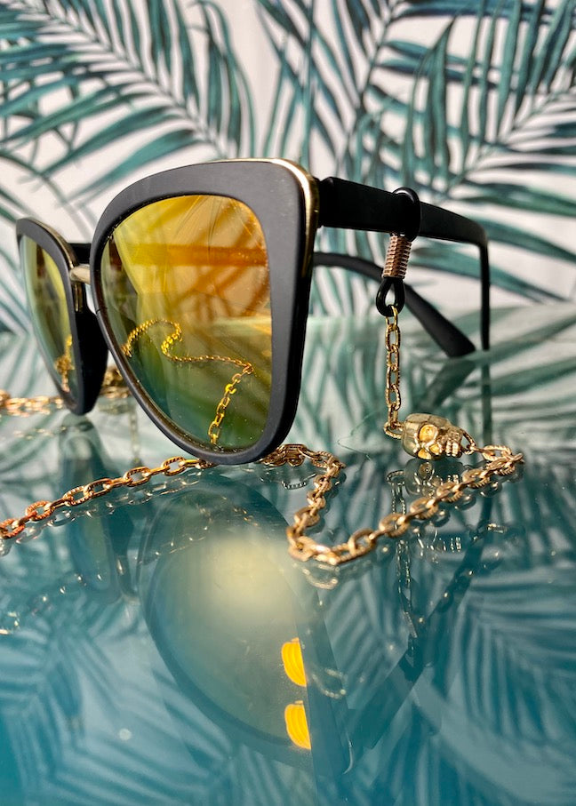 black sunglasses with gold skull chain on reflective table with palm background